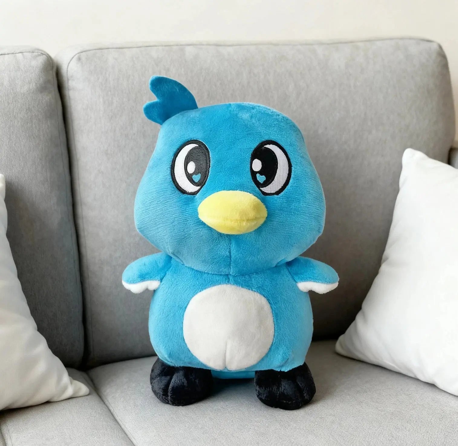 Blue and white plush bird toy with big eyes and yellow beak sitting on gray sofa with white pillows