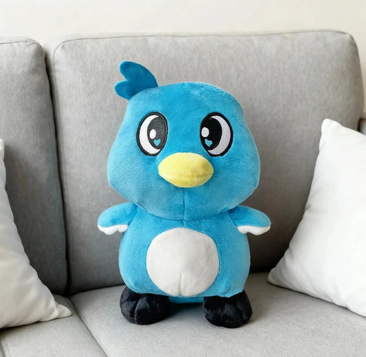 Blue and white plush bird toy with big eyes and yellow beak sitting on gray sofa with white pillows